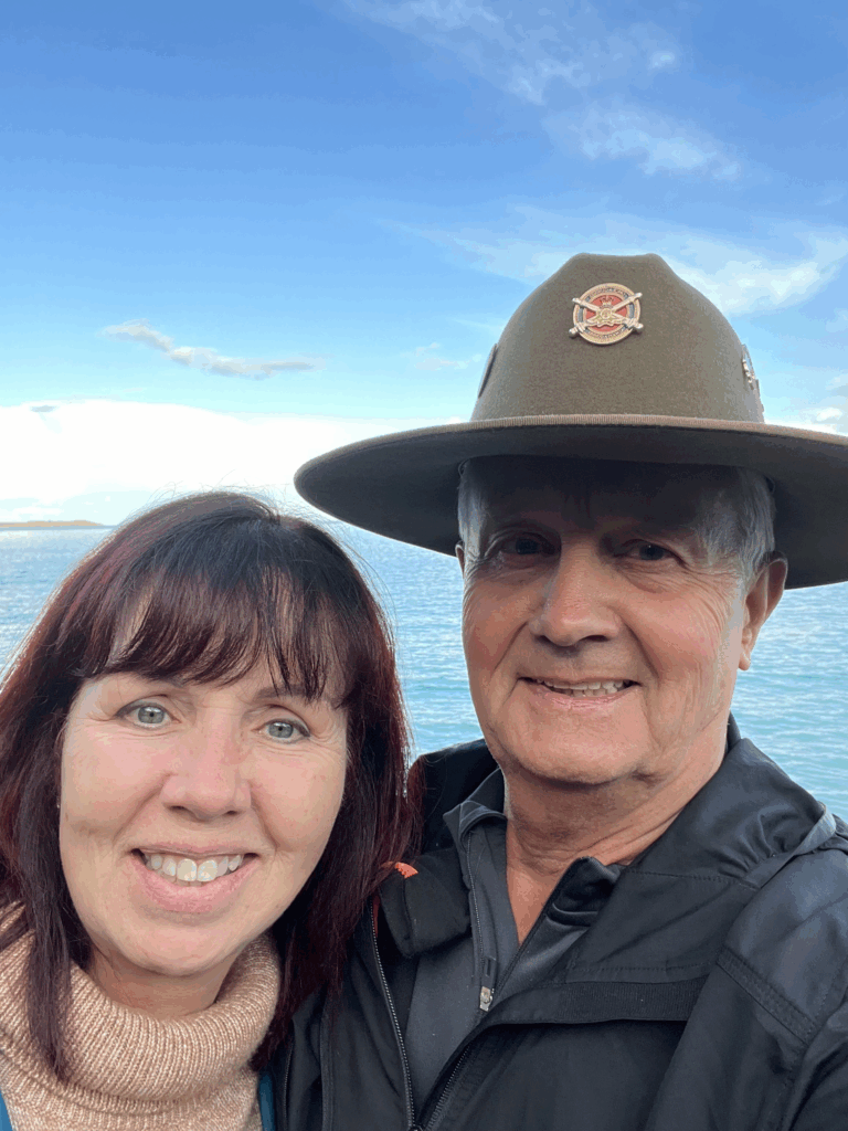 Smiling woman in a tan turtleneck beside a man wearing a wide-brimmed hat by a body of water and blue sky behind them.