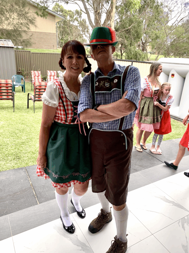 Couple in Bavarian costumes posing outdoors: woman in a red-and-white dirndl with green apron, man in lederhosen and a green hat with a red feather.